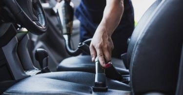 A person uses a small handheld vacuum to remove stains and debris from the driver's seat of a vehicle.