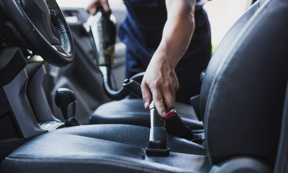 A person uses a small handheld vacuum to remove stains and debris from the driver's seat of a vehicle.