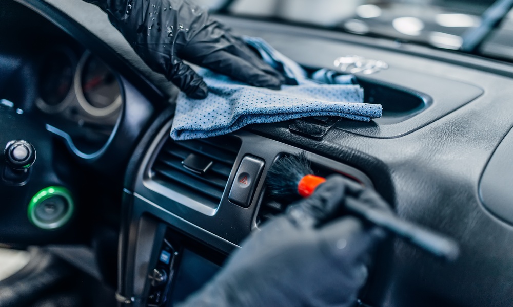 A person wearing black gloves uses a microfiber cloth to dust off a car's dashboard, while also brushing the air vent.