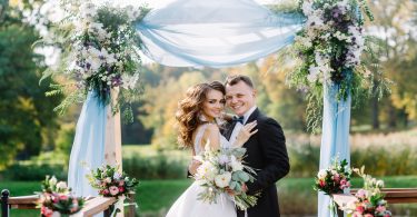 Smiling bride and groom embrace in front of a wedding altar decorated with vibrant flowers and soft blue gauze.