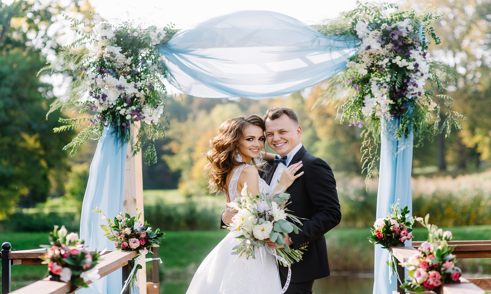 Smiling bride and groom embrace in front of a wedding altar decorated with vibrant flowers and soft blue gauze.