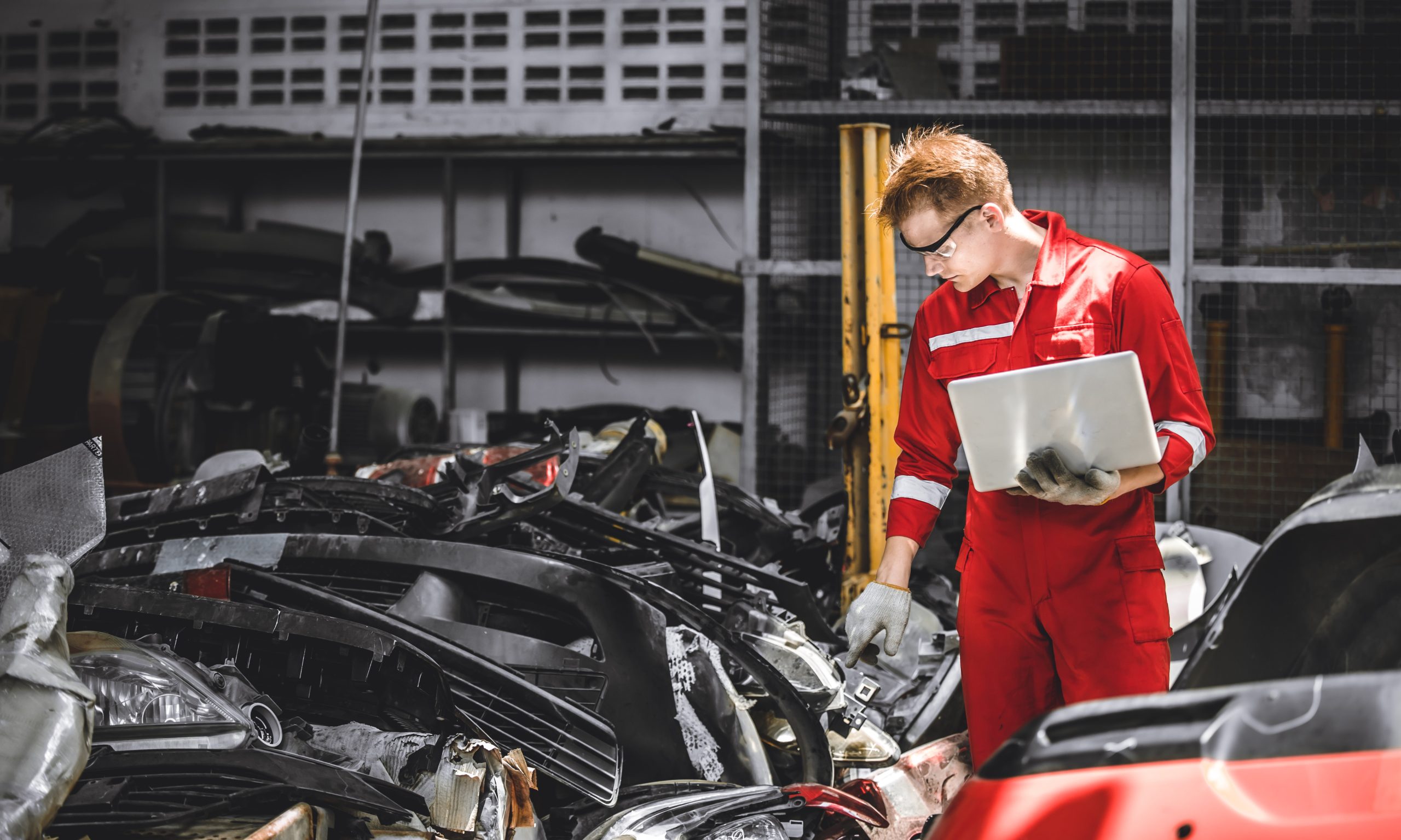 A worker looking over a pile of recycled parts from vehicles in a junkyard while holding a laptop in one hand.