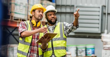 Two bearded men wearing plaid shirts, hi-vis vests, and hard hats are standing as they point to something.