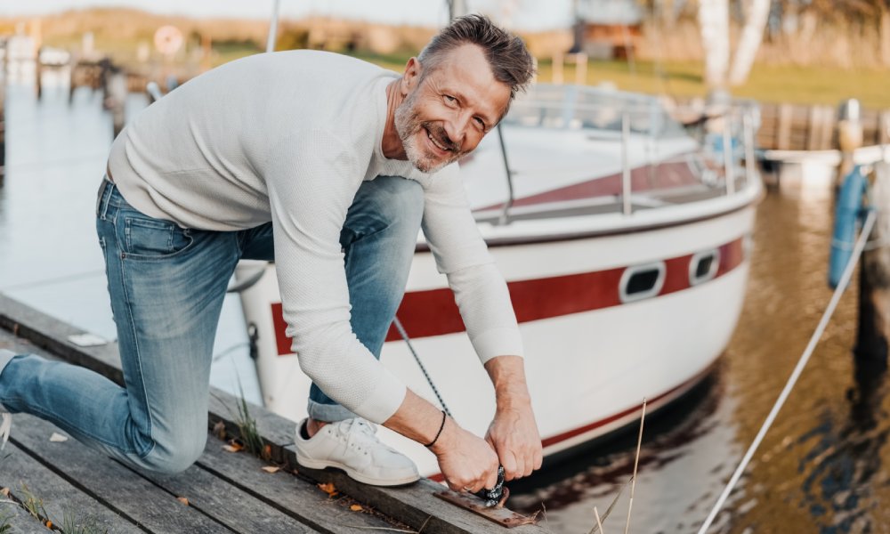 A close-up of a smiling, mature man tying up the mooring rope for his red sports yacht to an empty dock.
