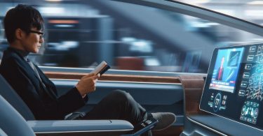 A passenger in an autonomous vehicle, reading a book as the car drives for him. He's in business casual attire.