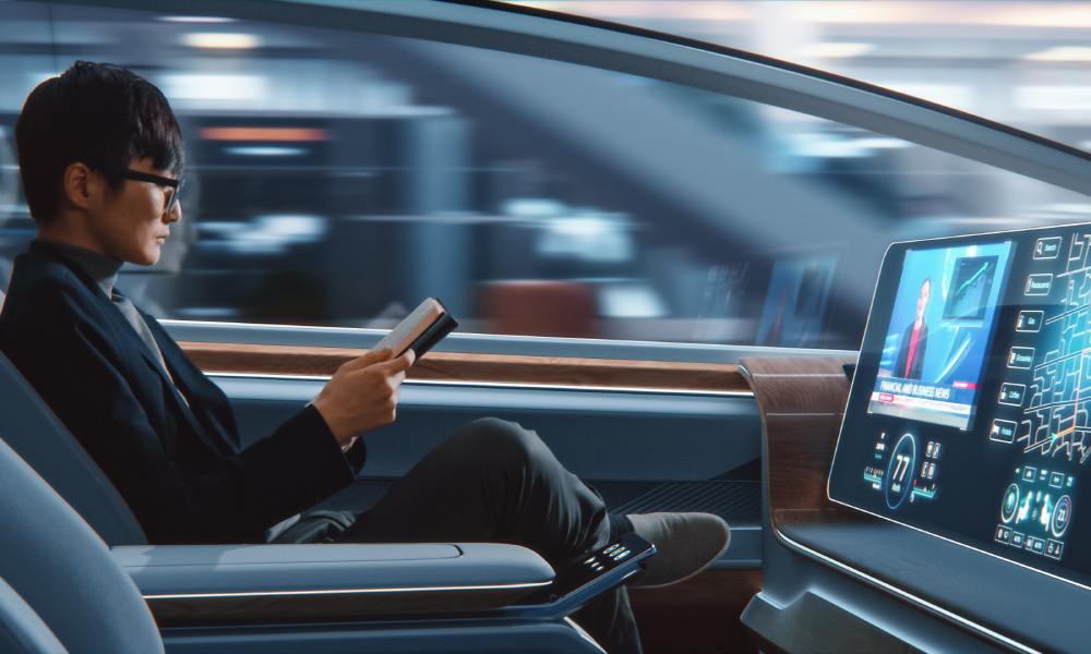 A passenger in an autonomous vehicle, reading a book as the car drives for him. He's in business casual attire.