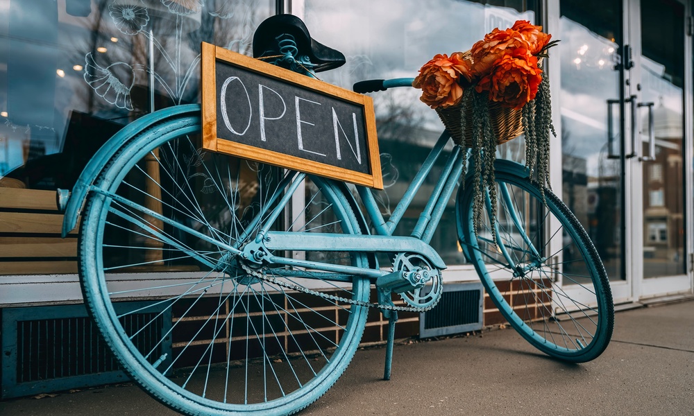 A blue bicycle in front of a store has a chalkboard open sigh hanging off of it and flowers in its basket.