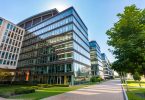 A modern office building with large glass windows. In front of the building is a long walking path with green trees.