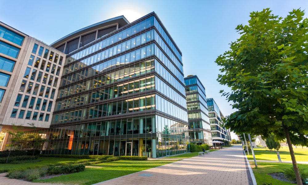 A modern office building with large glass windows. In front of the building is a long walking path with green trees.