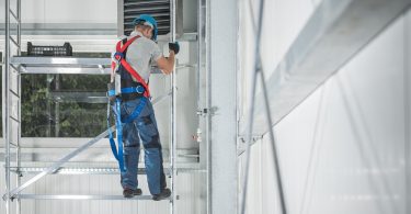 A construction worker in a harness, helmet, and overalls climbs metal scaffolding in a white warehouse.