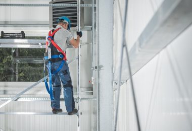 A construction worker in a harness, helmet, and overalls climbs metal scaffolding in a white warehouse.