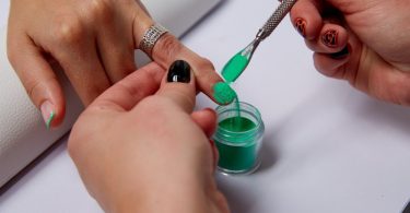 A nail technician holding a client's finger still above a small jar of green dip powder to catch excess during application.
