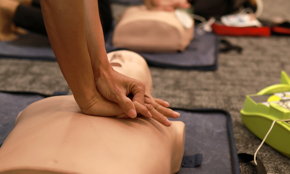 A close-up of a person practicing chest compressions on a mannequin during a class, with others in the background.