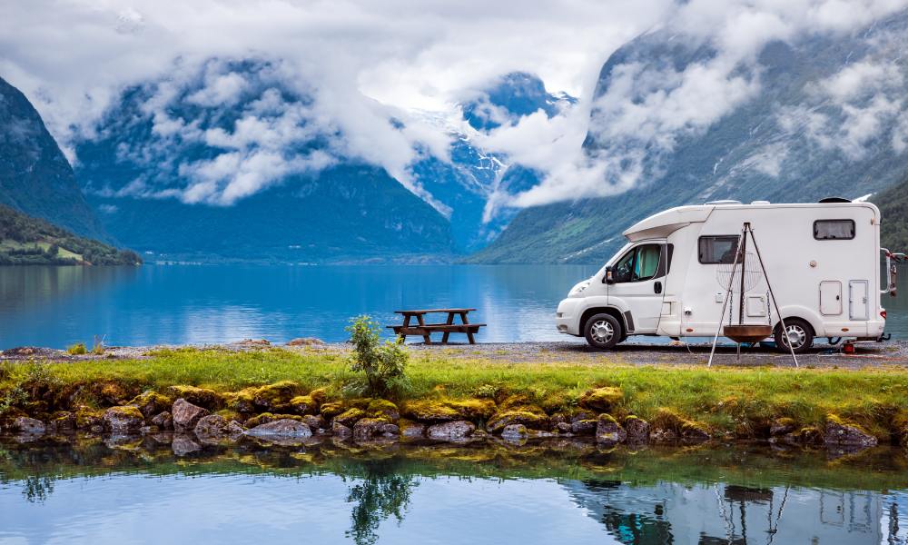 An RV is parked at the end of the road. In front is a picnic table and the view of a lake surrounded by mountains.
