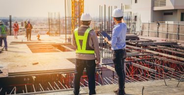 Two men in PPE stand overlooking a commercial construction site. One holds a tablet and points somewhere in the distance.