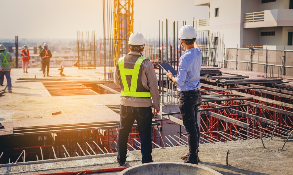 Two men in PPE stand overlooking a commercial construction site. One holds a tablet and points somewhere in the distance.