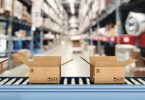 Cardboard boxes move on a conveyor belt in a warehouse with blurred shelving units stacked behind them.