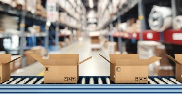 Cardboard boxes move on a conveyor belt in a warehouse with blurred shelving units stacked behind them.