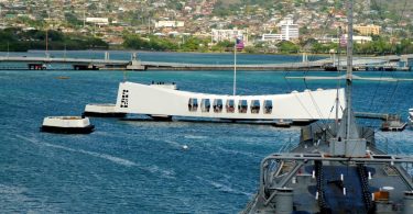The USS Arizona Memorial, a prominent, long white structure, topped with a flag, standing in the sea near a dock.