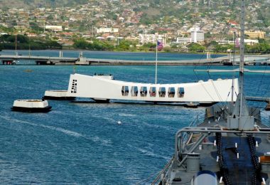The USS Arizona Memorial, a prominent, long white structure, topped with a flag, standing in the sea near a dock.