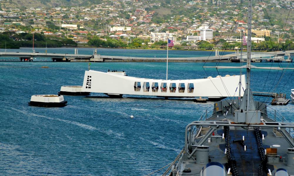 The USS Arizona Memorial, a prominent, long white structure, topped with a flag, standing in the sea near a dock.