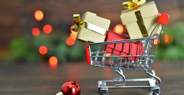 A miniature shopping cart filled with wrapped gift boxes on a wooden surface with festive lights behind it.