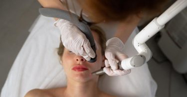 A woman in bold red lipstick lies on a medical bed as a beautician uses a medical laser on her face.