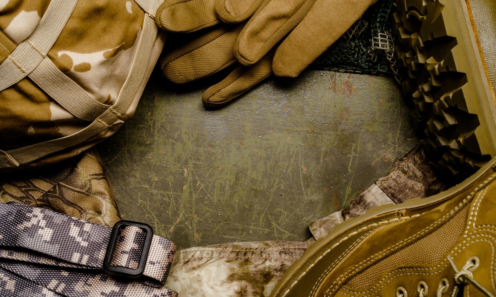 Military supplies on a scratched green table. The supplies include gloves, boots, a strap, and a bag.