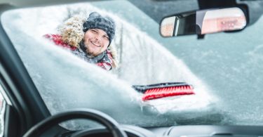 The interior of a car with a man wiping a pile of snow off the windshield with a snowbrush. He is wearing a winter coat.