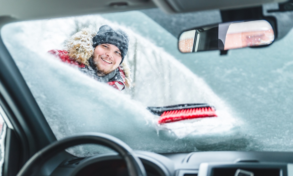 The interior of a car with a man wiping a pile of snow off the windshield with a snowbrush. He is wearing a winter coat.