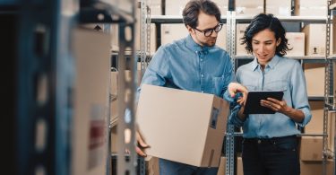 A man and a woman are standing next to each other. The woman is holding a tablet and they’re both looking at it.