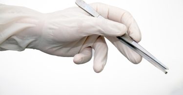 A latex-gloved hand grips shiny, slightly open metal forceps on a white background, evoking a sterile, clinical setting.