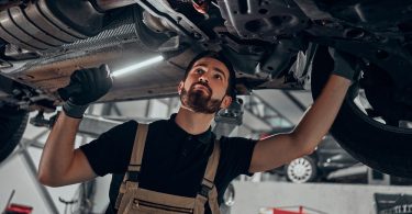 A mechanic wearing overalls stands below the undercarriage of an elevated car. He holds a small light.