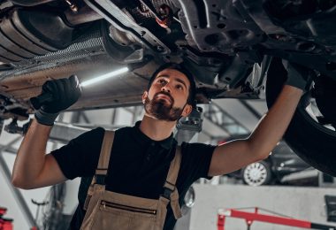 A mechanic wearing overalls stands below the undercarriage of an elevated car. He holds a small light.