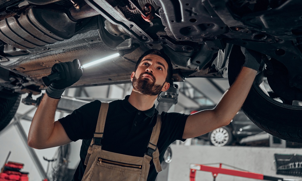 A mechanic wearing overalls stands below the undercarriage of an elevated car. He holds a small light.