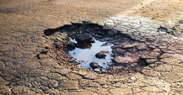 An asphalt lot has a giant pothole; the pothole itself has a puddle of water in it. There are faded white lines around the hole.