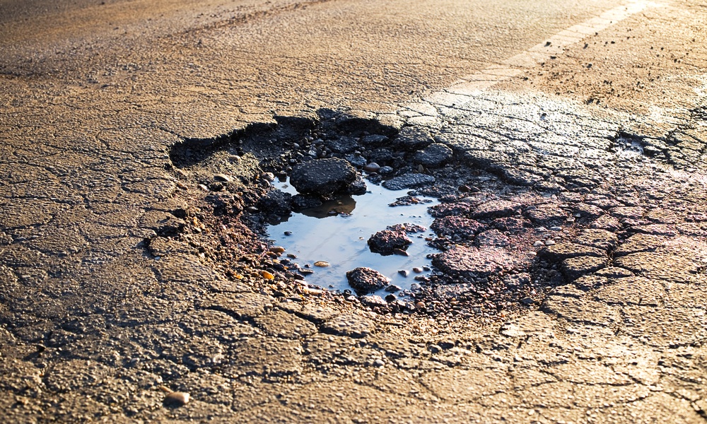 An asphalt lot has a giant pothole; the pothole itself has a puddle of water in it. There are faded white lines around the hole.
