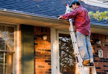A man standing on a ladder that leans against a house. He is cleaning the gutters on the roof with his hand.