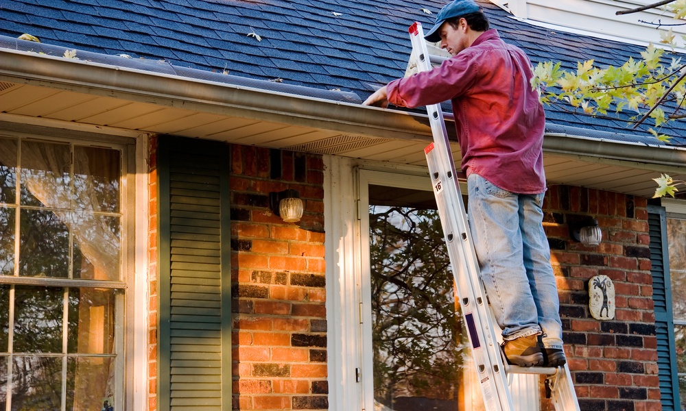 A man standing on a ladder that leans against a house. He is cleaning the gutters on the roof with his hand.