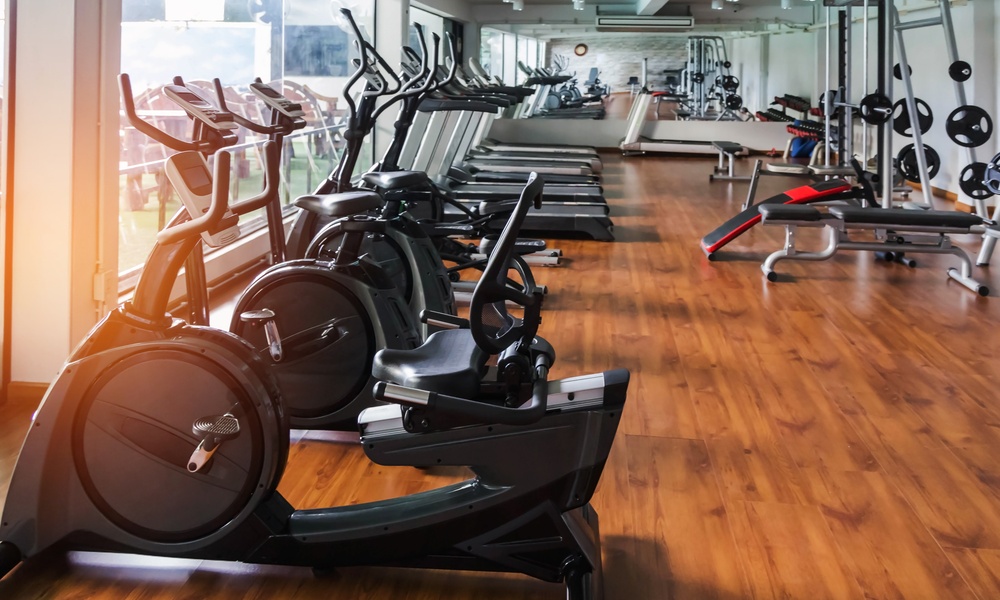 Several rows of stationary exercise bikes in a modern fitness center gym with strength equipment in the background.