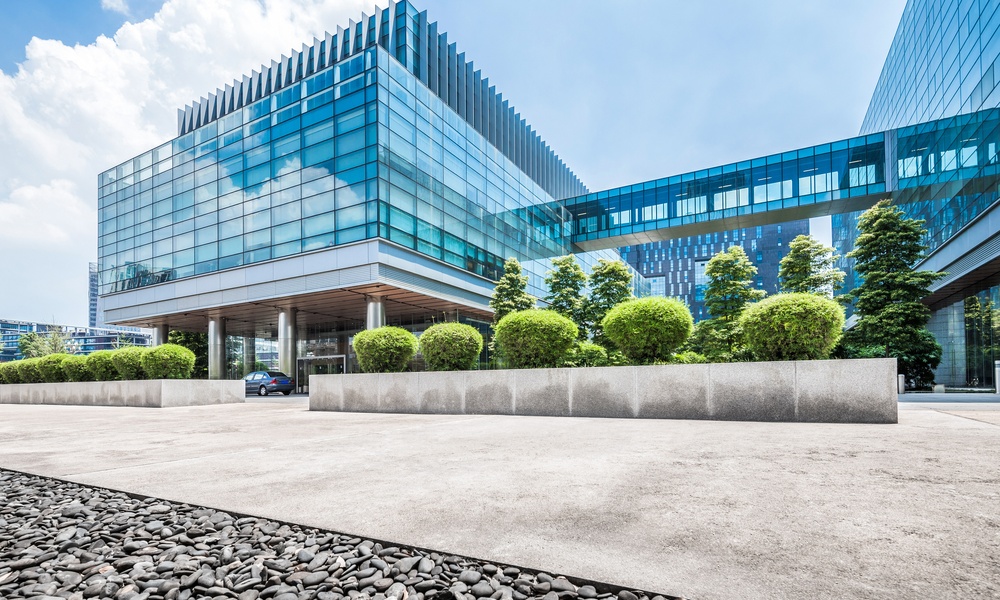 Modern glass buildings with a skywalk, lush greenery, and a paved foreground under a bright blue sky with scattered clouds.