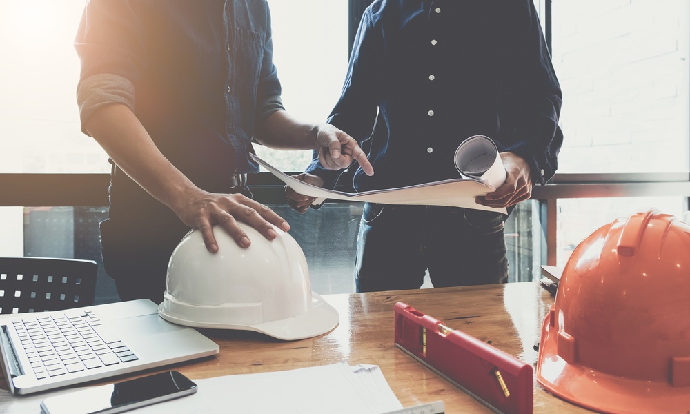Construction managers review building plans and blueprints at a table with hard hats, a laptop, and project documents.