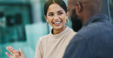 Two people are sitting in a glass office. The woman is smiling and speaking while holding a tablet, and the man listens.