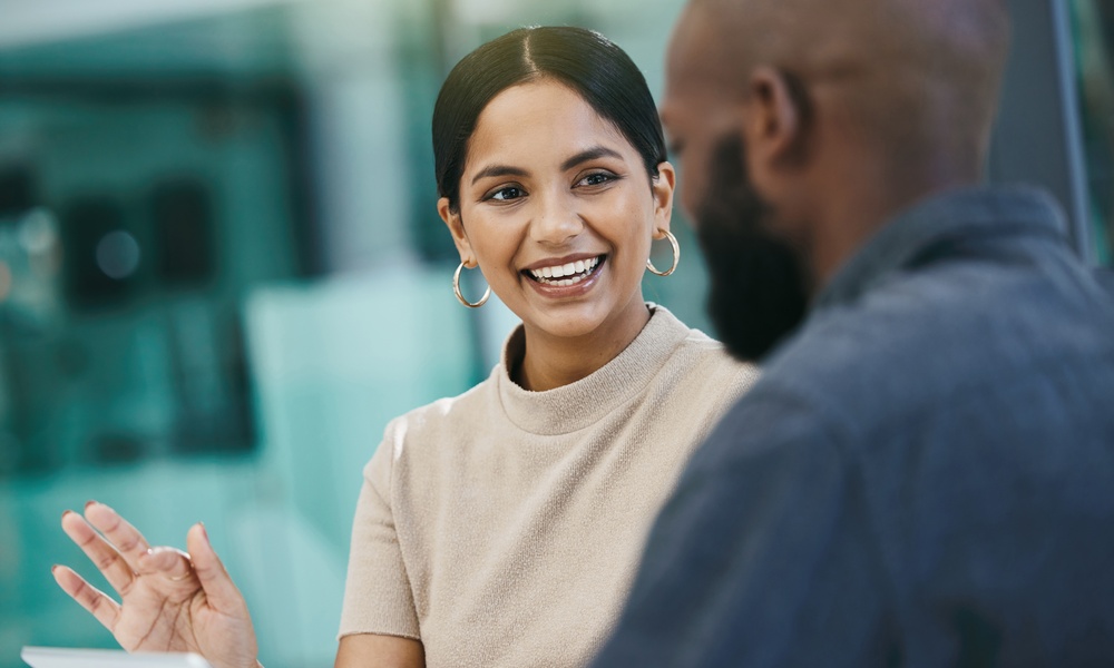 Two people are sitting in a glass office. The woman is smiling and speaking while holding a tablet, and the man listens.