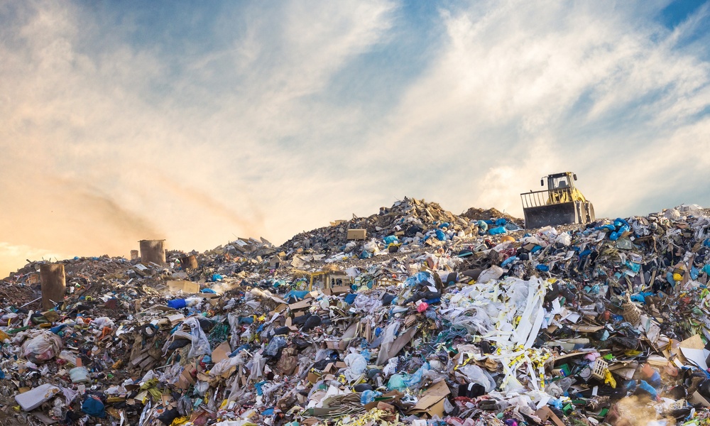Large landfill piled high with mixed garbage and plastic waste, bulldozer parked on top under cloudy sky.