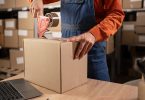 A person in an orange shirt and overalls holds a cardboard box in place on a table and tapes the top of the box.