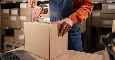 A person in an orange shirt and overalls holds a cardboard box in place on a table and tapes the top of the box.