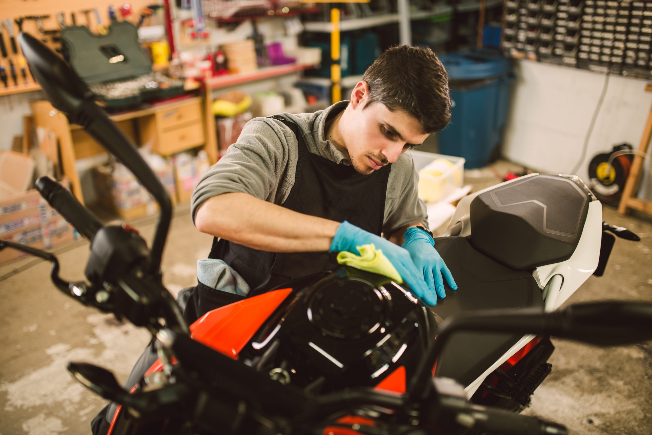 A man with short brown hair is kneeling next to a motorcycle as he cleans the body with a cloth.
