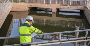 A professional engineer in a yellow safety vest standing above a large pool of water at a wastewater treatment plant.