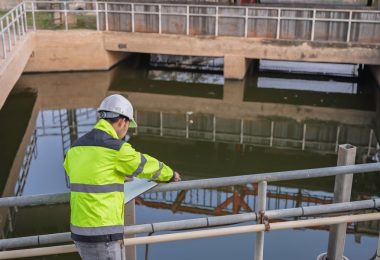 A professional engineer in a yellow safety vest standing above a large pool of water at a wastewater treatment plant.
